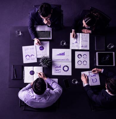 Vista aérea de uma reunião executiva, com profissionais reunidos ao redor de uma mesa analisando gráficos, relatórios e dados em notebooks.